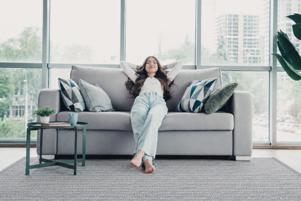 Rugmaker_Blog-Piece_Young woman leans back on a grey sofa in a bright modern apartment, enjoying a relaxed moment surrou